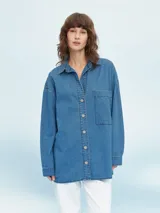 Camisa de denim azul de corte oversize, con mangas largas, cuello clásico, un bolsillo aplicado en el pecho y cierre frontal con botones metálicos.