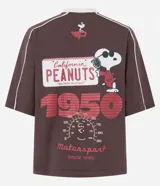 Remera de algodón color marrón con cuello redondo y mangas cortas. Presenta un estampado frontal con el personaje Snoopy en temática de carreras y detalles de líneas blancas contrastantes en los hombros y mangas.