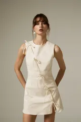 Vestido corto de jean color natural, con corte al cuerpo, cuello a la base y diseño sin mangas. Presenta un detalle de cierre frontal con lazos decorativos y una pequeña abertura lateral.