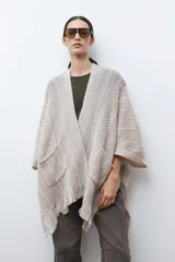 Ruana de tejido de punto en tonos beige y blanco, con patrón de espiga. Presenta cuello en V, mangas amplias, dos bolsillos frontales y flecos en el dobladillo.
