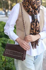 Cartera tipo sobre color marrón chocolate, confeccionada en cuero texturizado con detalle de tachas metálicas plateadas en la solapa frontal. Incluye una correa de cadena metálica con eslabones entrelazados y cierre principal con cremallera.