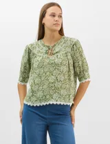 Blusa de gasa estampada con motivos botánicos en color verde, con escote redondo, abertura frontal con lazos de ajuste y mangas cortas. Presenta detalles de encaje en el escote, mangas y ruedo.