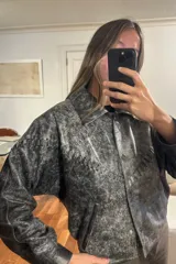 Campera bomber de cuero ovino con efecto desgastado en tonos grisáceos y negros. Presenta cuello camisero, cierre frontal con broches forrados y diseño holgado.