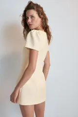 Vestido mono corto de color beige claro, con cuello redondo y mangas cortas abullonadas. Cuenta con forro interior tipo shorts y cierre en la espalda con cremallera oculta.