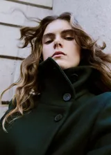 Abrigo largo de paño en color verde oscuro, con cuello alto, cierre frontal oculto y cinturón ajustable del mismo tejido. Presenta un diseño estructurado con bolsillos laterales.