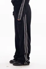 Pantalón deportivo negro de calce relajado con cintura elástica. Presenta una franja lateral gruesa de color gris claro con bordes blancos. Se observa un pequeño bordado en la zona del bolsillo con la palabra "Delosalto".