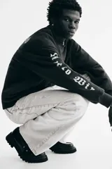 Buzo negro de algodón de peso medio con cuello redondo, hombros caídos y remates acanalados. Presenta un estampado frontal de una calavera rodeada de texto y texto en ambas mangas en tipografía gótica.