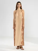 Vestido largo tipo maxi con cuello halter, sin mangas y corte holgado. Presenta un diseño estampado de flores en tonos marrones sobre una base de color beige.