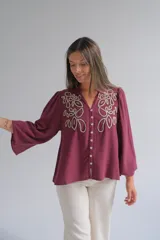Blusa color bordó con bordados florales en tono crema en la parte superior frontal. Presenta cuello redondo con pequeña abertura, cierre frontal con botones y mangas largas acampanadas con puños ajustados.