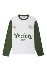 Remera de manga larga con diseño estilo camiseta de fútbol, cuerpo blanco y mangas verdes. Presenta estampados gráficos en el frente con la inscripción 'Victory Legend', el número '39' y un emblema ovalado.