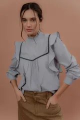 Blusa de jeans fino color celeste con detalles de terciopelo negro en contraste. Presenta cuello alto con volados, mangas largas abullonadas con puños ajustados y volados decorativos en los hombros y el frente.