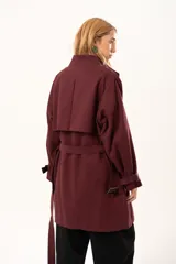 Trench de corte holgado color bordó, con cuello camisero, cierre frontal con botones y cinturón ajustable con hebilla. Presenta mangas largas con detalles de correas en los puños y bolsillos laterales.