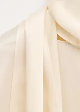 Blusa de seda color blanco marfil, con cuello alto drapeado y detalle de bufanda integrada al cuello. De manga larga con puños ajustados mediante tiras.