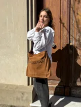 Cartera tipo tote bag de cuero color camel, con detalles de tachas metálicas en el bolsillo frontal y tiras largas para el hombro.