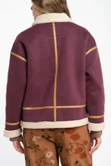 Campera tipo gamulán de color bordó, con interior y cuello de corderito sintético en tono crema. Presenta detalles de ribetes en cuero sintético color suela, cierre frontal metálico y bolsillos laterales.