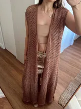 Chaleco largo sin mangas, tejido en punto calado tipo red con hilo de algodón y lurex, color marrón. Presenta escote profundo en V, tiras para atar en el frente, tajo trasero y flecos en el ruedo.