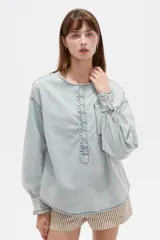 Blusa de jean ligera en color celeste claro, con cuello redondo y cierre frontal parcial con botones. Presenta un diseño con volados a lo largo de la botonera, bolsillo ojal en el pecho, y mangas largas abullonadas con puños ajustados y fruncidos.