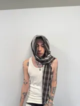 Bufanda larga con diseño de cuadrille en tonos marrón, negro y blanco. Presenta flecos en los extremos, una grifa de goma con logo en una punta y una etiqueta tejida con el nombre de la marca en la otra.