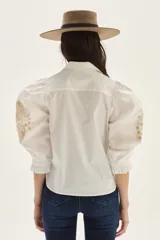 Camisa blanca de algodón con mangas abullonadas y puños fruncidos. Presenta bordados en tono beige/tostado a lo largo de la botonera frontal y un diseño bordado grande en forma de hoja o pluma en la manga.