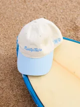 Gorra de lona color crema con visera celeste. Presenta bordado frontal en hilo azul claro con el texto "Rusty Tribe" y un pequeño diseño de flores.