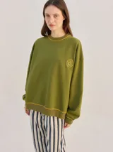 Buzo oversize color verde militar, de corte crew con cuello redondo, mangas largas y puños ajustados. Cuenta con detalles de costura en zigzag amarilla en cuello, puños y ruedo, y un pequeño logo bordado en el pecho.