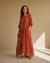Vestido largo color terracota de lino, con diseño asimétrico que incluye un panel lateral estampado con grandes flores oscuras, escote redondo, mangas 3/4 con trabilla y botón para arremangar, y un amplio bolsillo frontal aplicado en el pecho con terminaciones deshilachadas.