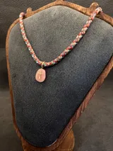 Collar de diseño artesanal compuesto por cordones trenzados multicolor en tonos verdes, rosados y rojizos. Presenta un dije de piedra ovalada con una cruz dorada aplicada en el centro.