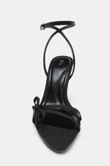 Sandalia de tacón alto color negro, con acabado satinado. Presenta tiras finas con un lazo decorativo en la parte delantera y cierre de hebilla en la pulsera del tobillo. La punta es redonda y el tacón mide 10 cm.