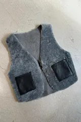 Chaleco reversible confeccionado en cuero genuino. Presenta un lado de piel de oveja gris con bolsillos de cuero negro y un lado de cuero liso gris. Diseño sin mangas con cierre frontal.