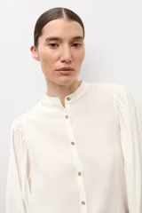 Camisa blanca de corte recto con cuello mao, cierre frontal de botones y mangas largas abullonadas con puños ajustados.