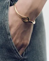 Pulsera rígida tipo brazalete PDPAOLA modelo Brise, chapada en oro, con un diseño fluido y entrelazado que forma un nudo central.