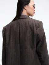 Blazer de corte sastrero color marrón con diseño de rayas verticales finas. Presenta solapas clásicas, cierre frontal de doble botonadura y un detalle de cadena metálica dorada en el bolsillo superior.