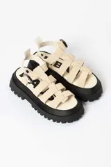 Sandalia tipo gladiadora con plataforma track negra. Presenta un diseño bicolor con tiras delanteras en color amarillo neón y talón con tira ajustable en color beige. Incluye hebilla metálica negra.