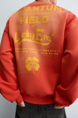 Sudadera de corte relajado, color naranja con efecto tie-dye/deslavado, cuello redondo y manga larga. Presenta un estampado frontal con texto y un trébol de cuatro hojas en amarillo.