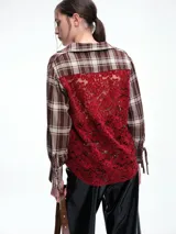 Camisa de corte relajado con estampado de cuadros en tonos negro y beige. Presenta cuello camisero, cierre frontal parcial con botones y mangas largas con puños ajustables mediante lazos. El diseño destaca por un detalle de encaje en la espalda que aporta contraste.