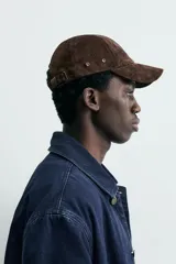 Gorra de béisbol color marrón oscuro, confeccionada en material con textura de ante o nobuk. Presenta visera curva y ojales metálicos a los lados para ventilación.