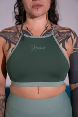 Top deportivo verde oscuro de doble tela, con cuello halter, espalda cruzada y banda elástica inferior en tono más claro. Cuenta con el logo de la marca estampado en el centro del pecho.