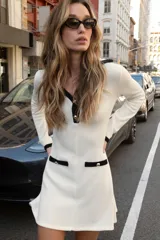 Vestido corto de tejido suave, color negro con detalles en blanco. Presenta cuello solapa, escote en pico, manga larga, falsos bolsillos delanteros y cierre frontal con botones dorados.
