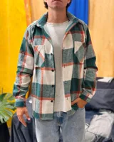 Sobrecamisa de paño con diseño a cuadros en tonos verde, blanco y detalles en naranja. Presenta cuello clásico, cierre frontal con botones, mangas largas con puños abotonados y dos bolsillos frontales con solapa en el pecho.