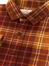 Camisa de manga larga confeccionada en franela de algodón, con diseño de cuadros en tonos marrones, mostaza y blanco. Presenta cuello con botones, cierre frontal abotonado y bolsillo en el pecho.