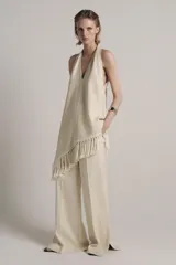 Top halter color crudo, de escote en V y diseño confeccionado en hilatura con mezcla de viscosa. Presenta bajo asimétrico con flecos al tono y cierre en la espalda.