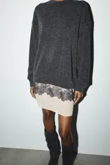 Vestido de cuello redondo y manga larga, confeccionado en tejido de punto gris oscuro. Presenta un bajo en contraste de color beige con detalle de encaje floral negro y lazada lateral ajustable.