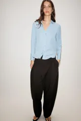 Camisa de cuello solapa y escote en pico, con manga larga y cierre frontal mediante botones. Presenta un diseño fluido con bajo asimétrico y aberturas laterales.