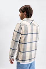 Sobrecamisa de tejido tipo polar con estampado de cuadros en tonos beige, gris y azul. Presenta cuello clásico, cierre frontal con botones oscuros y bolsillos frontales en el pecho.