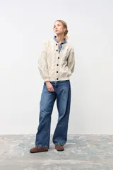 Cardigan de punto grueso color crudo, con diseño de trenzas verticales, cuello redondo y cierre frontal con botones oscuros.