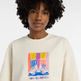 Remera corta de manga corta, color beige claro, con cuello redondo. Presenta un estampado frontal colorido que muestra un par de zapatillas con calcetines a rayas de colores vibrantes (amarillo, fucsia, azul) y el texto "new balance" debajo.