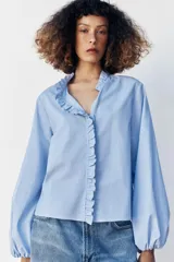 Blusa celeste de popelín, con cuello subido y escote de pico, manga larga abullonada terminada en elástico, y detalles de volantes en el cuello y la botonadura frontal con cierre de botones ocultos.