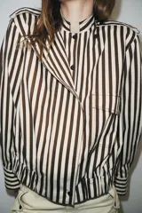 Camisa tipo bomber confeccionada en algodón, con diseño de rayas verticales en tonos marrón y blanco. Presenta cuello perkins, manga larga con puños, bolsillo de plastrón con solapa en el frente, bajo elástico y cierre frontal con botones.