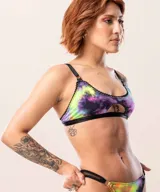 Corpiño de estilo deportivo con estampado tipo galaxia en tonos violeta, rosa y verde neón. Presenta un diseño con abertura frontal, bordes con detalle de puntilla negra y elástico negro en la base. Cuenta con breteles regulables y aros metálicos en la unión de los breteles.