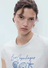 Remera de manga corta color blanco con cuello redondo y diseño recto. Presenta un estampado frontal en tonos azules con ilustraciones de caracoles y la inscripción "Les Coquillages".